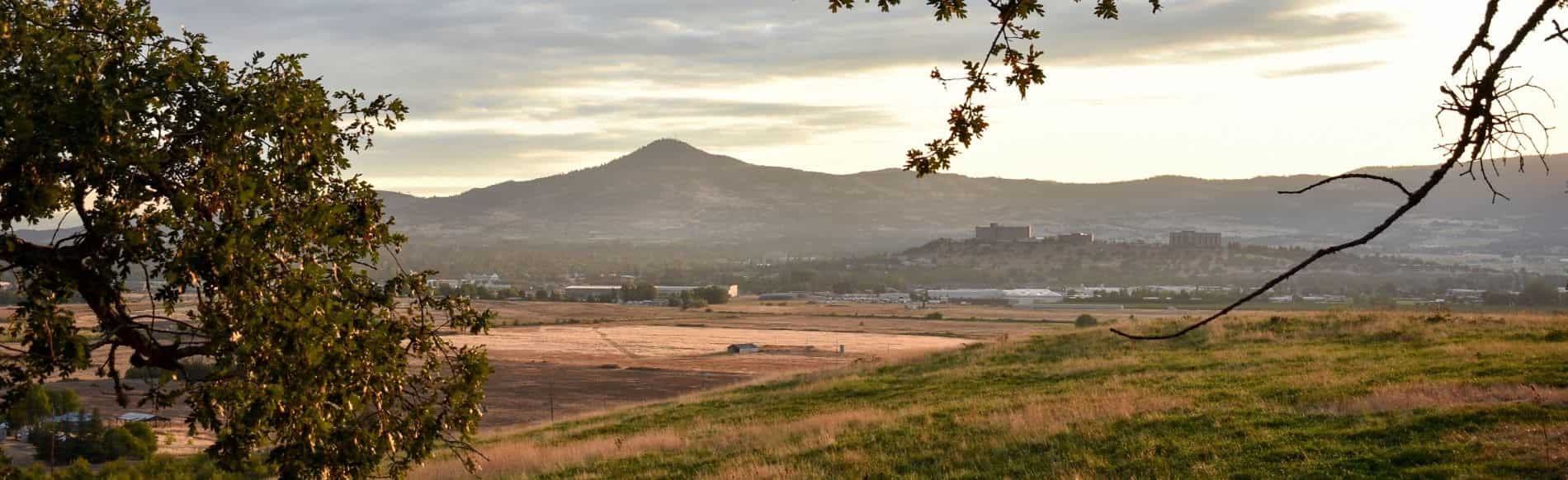 View of the Rogue Valley from a hillside near Medford, Oregon, with Table Rock and distant buildings under a hazy sunset sky framed by oak branches